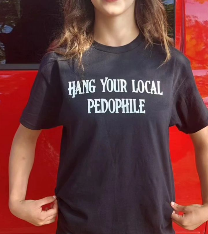 Young person with long hair wearing a shirt that says "Hang Your Local Pedophile" standing in front of a vehicle.